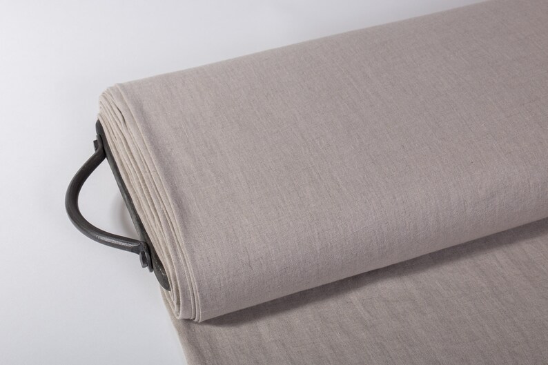 May include: A roll of light gray fabric, possibly linen or a similar textile, is displayed. The fabric is rolled on a dark metal holder with a curved handle. The material has a slightly textured appearance.