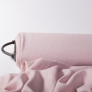 May include: A roll of light pink fabric with a visible woven texture, wrapped around a dark metal cylindrical core featuring a curved handle on the side. A portion of the soft, draped fabric extends outward from the main roll onto the surface.