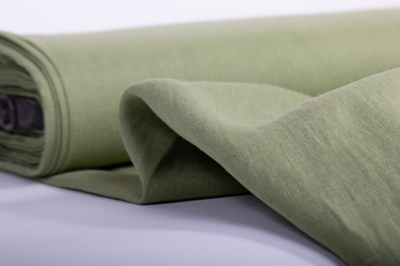May include: A roll of soft, green linen fabric. The fabric is folded and draped, showing its texture and drape.