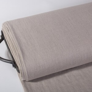 Puede incluir: Un rollo de tela de lino beige claro en un estante de metal con un asa. La tela tiene una textura sutil y est&aacute; lista para ser utilizada para coser o proyectos de manualidades.