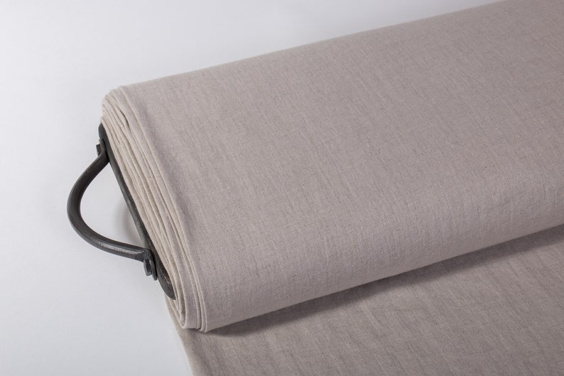 Puede incluir: Un rollo de tela de lino beige claro en un estante de metal con un asa. La tela tiene una textura sutil y est&aacute; lista para ser utilizada para coser o proyectos de manualidades.