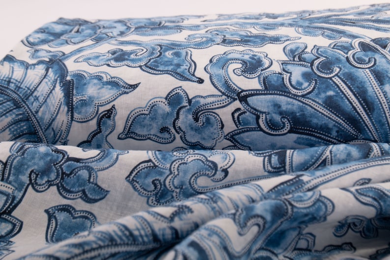 May include: Close-up of folded fabric with a repeating blue and white pattern. The design features stylized cloud and floral motifs. The fabric appears to be linen or a similar natural fibre, with a slightly textured surface.