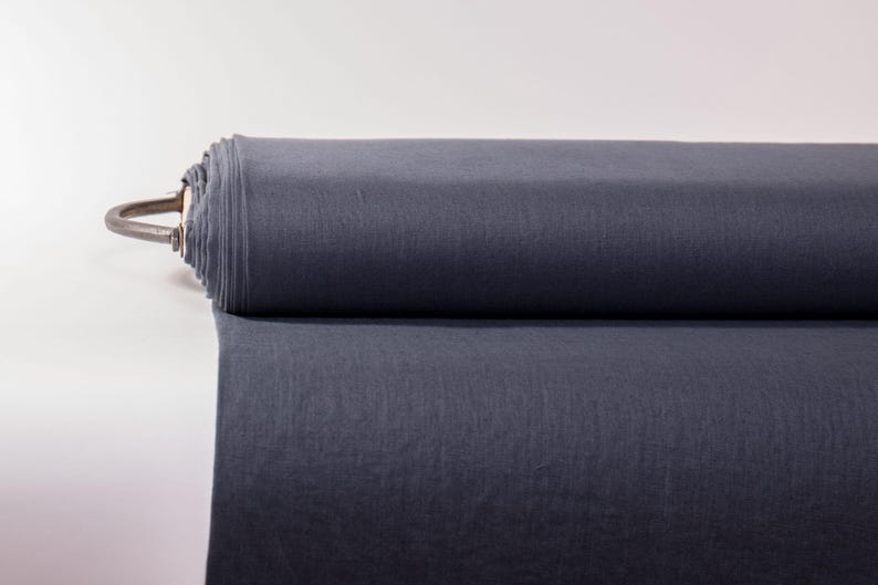 May include: A large roll of solid dark charcoal gray fabric. The textile is tightly wound around a central core, with a portion of the material unrolled and extending forward, showcasing a smooth, matte surface and a consistent, deep gray color.