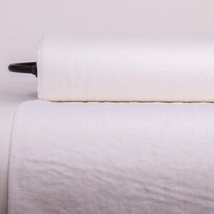 May include: A large roll of solid white fabric with a visible woven texture. The cylindrical roll is positioned horizontally, with a black plastic end cap visible on the left side, and a length of the same white fabric draped downward in front.