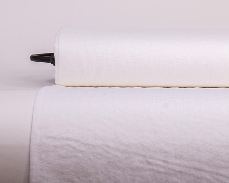 May include: A large roll of solid white fabric with a visible woven texture. The cylindrical roll is positioned horizontally, with a black plastic end cap visible on the left side, and a length of the same white fabric draped downward in front.
