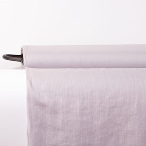 May include: A roll of light lavender fabric featuring a visible woven texture. The fabric is wrapped around a dark, cylindrical core that extends slightly from the side of the roll, showcasing the material's soft, muted color and consistent weave.