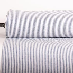 May include: A roll of fabric featuring a vertical pinstripe pattern in blue and white. The textile is tightly wound around a central dark-colored cylindrical core, displaying a consistent, repeating linear design across the entire visible surface area.
