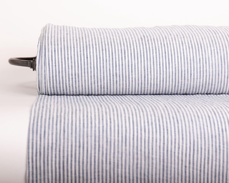 May include: A roll of fabric featuring a vertical pinstripe pattern in blue and white. The textile is tightly wound around a central dark-colored cylindrical core, displaying a consistent, repeating linear design across the entire visible surface area.