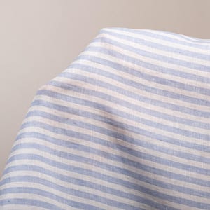 May include: A piece of fabric featuring a pattern of horizontal light blue and white stripes. The textile has a visible woven texture and is draped to show folds and depth, highlighting the consistent alternating color scheme across the surface.