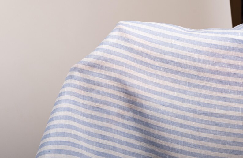 May include: A piece of fabric featuring a pattern of horizontal light blue and white stripes. The textile has a visible woven texture and is draped to show folds and depth, highlighting the consistent alternating color scheme across the surface.