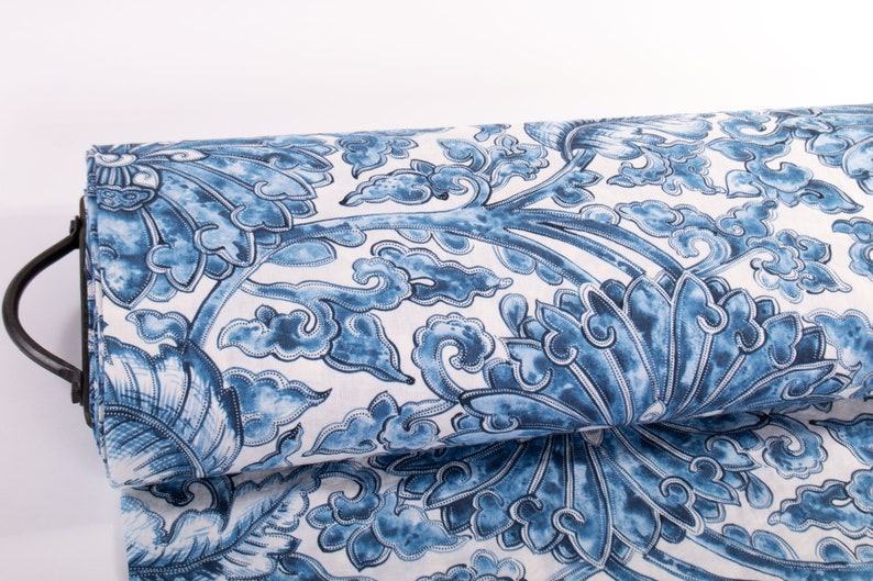 May include: A roll of fabric displaying a blue and white floral design. The pattern includes stylized flowers, leaves, and cloud motifs. The fabric is held by a black metal rod, indicating it's a bolt of material suitable for sewing or crafts.