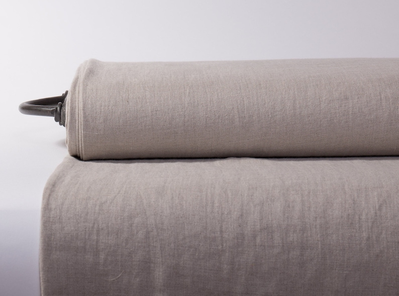 May include: A roll of natural-colored linen fabric. The fabric is a light beige color and appears to be tightly rolled. The texture of the linen is visible, suggesting a high-quality material. A metal rod is attached to the roll.