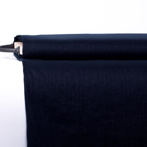 May include: A bolt of solid dark navy blue fabric wrapped around a wooden core. The textile features a visible woven texture and a matte finish, with a portion of the fabric draped downward from the horizontal roll against a plain white background.