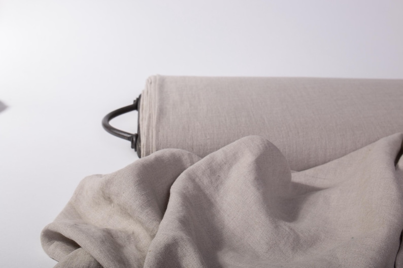 May include: A roll of light beige fabric with a visible woven texture, featuring a dark metal handle attached to the side. A portion of the fabric is unrolled and draped in front, showcasing the soft, natural appearance of the material.