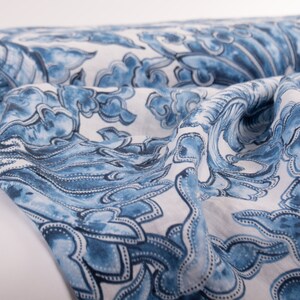 May include: A roll of fabric with a blue and white floral pattern. The design features stylized flowers and cloud-like shapes in varying shades of blue against a white background. The fabric is folded and draped, showcasing its texture.