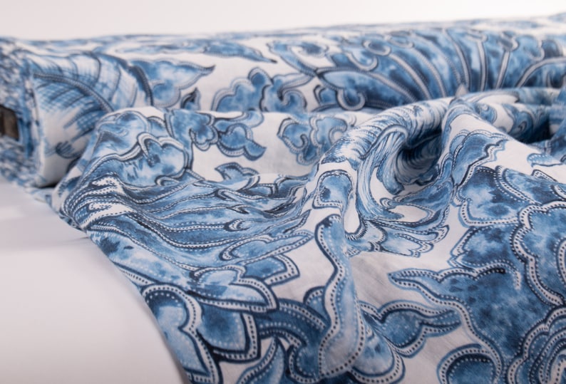 May include: A roll of fabric with a blue and white floral pattern. The design features stylized flowers and cloud-like shapes in varying shades of blue against a white background. The fabric is folded and draped, showcasing its texture.