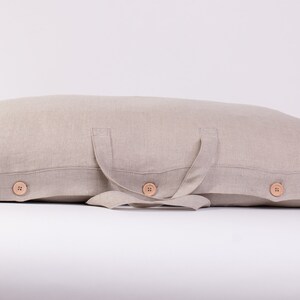 Puede incluir: Una almohada de lino beige con dos asas de tela y tres botones de madera. La almohada est&aacute; doblada por la mitad y las asas est&aacute;n atadas juntas en un nudo.