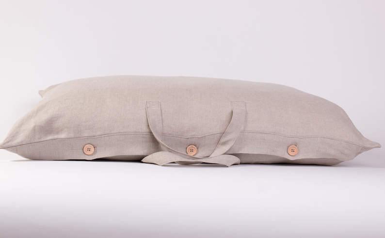 Puede incluir: Una almohada de lino beige con dos asas de tela y tres botones de madera. La almohada est&aacute; doblada por la mitad y las asas est&aacute;n atadas juntas en un nudo.