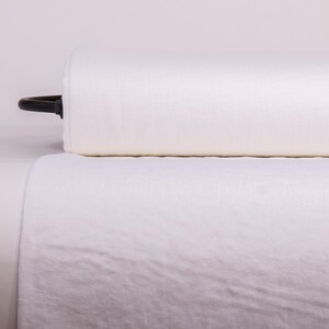May include: A large roll of off-white fabric featuring a smooth, tightly woven texture. The cylindrical bolt of material is positioned horizontally, with a small black plastic end cap visible on the left side of the roll, showing a clean finish.
