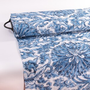 May include: A roll of fabric with a blue and white floral design. The pattern showcases large stylized flowers and swirling cloud-like forms. The fabric is rolled on a black metal rod.