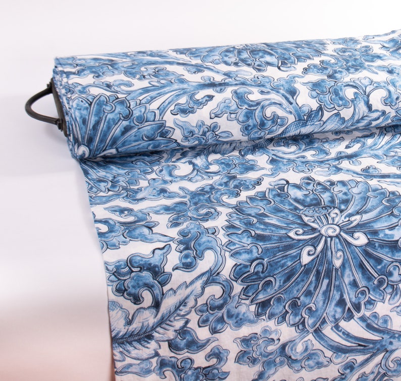 May include: A roll of fabric with a blue and white floral design. The pattern showcases large stylized flowers and swirling cloud-like forms. The fabric is rolled on a black metal rod.