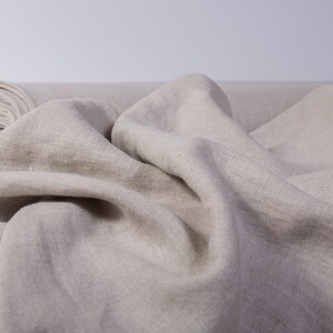 May include: A roll of natural-colored linen fabric, partially unrolled, with soft folds and a textured surface. The fabric is a neutral beige tone, suitable for various textile projects. The roll is visible in the background.