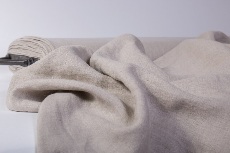 May include: A roll of natural-colored linen fabric, partially unrolled, with soft folds and a textured surface. The fabric is a neutral beige tone, suitable for various textile projects. The roll is visible in the background.