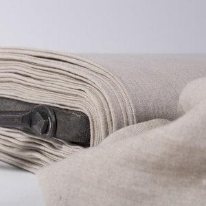 Puede incluir: Un rollo de tela de lino natural, doblado y apilado en un carrete de madera con un mango de metal. La tela es de color beige claro y tiene una superficie suave y texturizada.