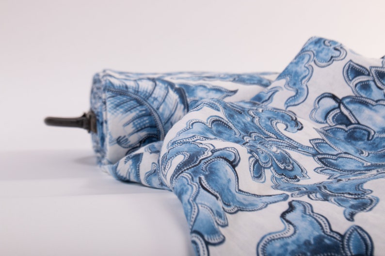 May include: A roll of fabric featuring a blue and white floral design. The fabric is unrolled, displaying a detailed pattern of stylized flowers and leaves. The roll is held by a dark brown rod.