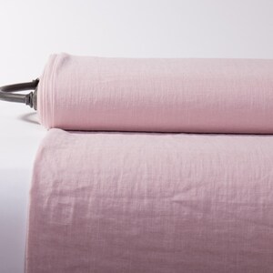 May include: A large roll of light pink fabric featuring a visible woven texture. The fabric is wrapped around a dark metal core that extends from the left side of the roll, showcasing a smooth, consistent color throughout the entire length.