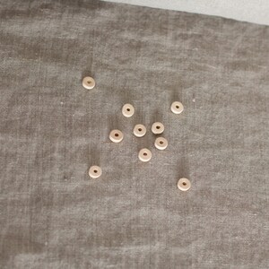May include: A collection of small, light-colored wooden beads with a hole in the center, scattered on a textured, neutral-toned fabric. The beads appear to be unfinished and natural in color, suitable for crafting or jewelry making.