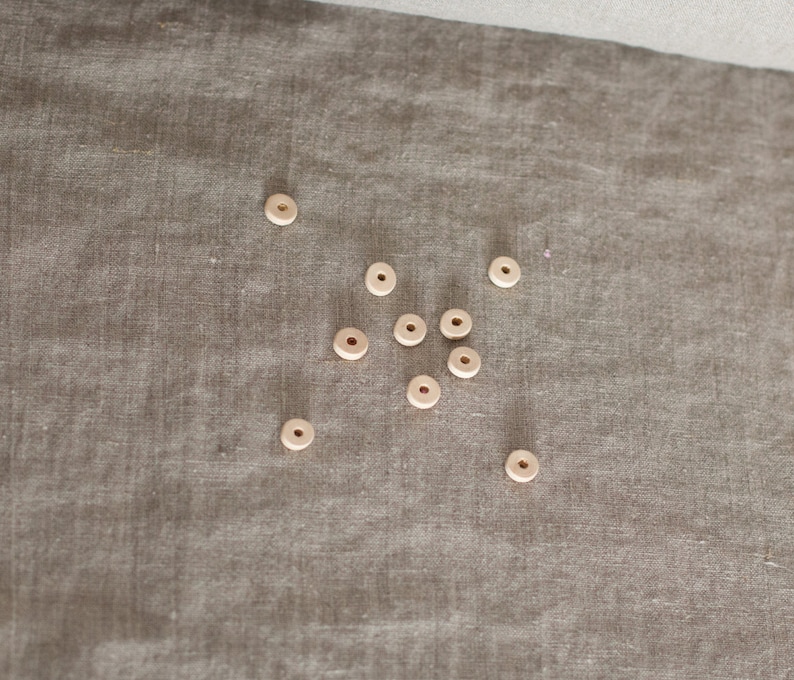 May include: A collection of small, light-colored wooden beads with a hole in the center, scattered on a textured, neutral-toned fabric. The beads appear to be unfinished and natural in color, suitable for crafting or jewelry making.