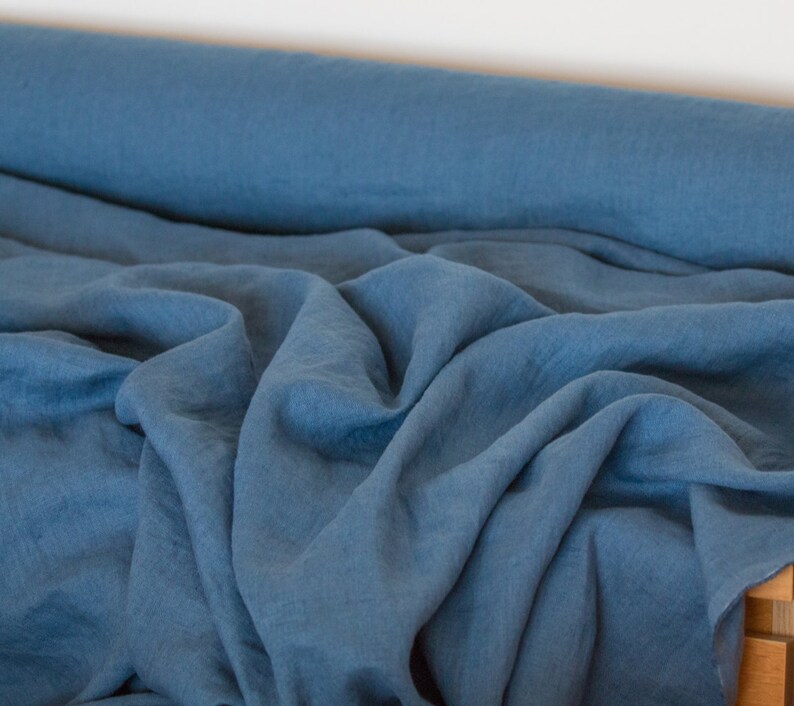May include: A roll of blue linen fabric with a soft, textured surface. The fabric is folded and draped, showcasing its natural wrinkles and drape.