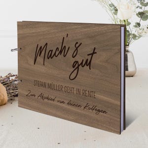 May include: A wooden photo album with German text including "Mach's gut". The album is brown with a wood grain effect and metal ring binding. It rests on a white surface, with flowers in the background. The album is a retirement gift.