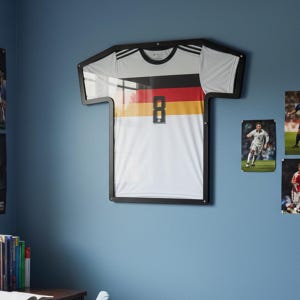 Könnte beinhalten: Gerahmtes deutsches Fußballtrikot mit der Nummer 8, an einer blauen Wand. Das Trikot ist weiß mit schwarzen Akzenten und den Farben der deutschen Flagge. Fußballposter sind ebenfalls an der Wand zu sehen.