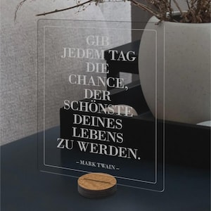 Op de afbeelding: Een helder acryl bord met de quote "GIB JEDEM TAG DIE CHANCE, DER SCHÖNSTE DEINES LEBENS ZU WERDEN." van Mark Twain, ondersteund door een houten basis. De tekst is gegraveerd in een modern lettertype.