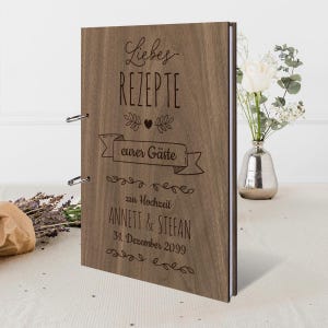 Könnte beinhalten: Ein hölzernes Rezeptbuch mit dem Text "Liebes Rezepte eurer Gäste zur Hochzeit ANNETT & STEFAN 31. Dezember 2099" auf dem Einband. Das Buch ist braun mit silbernen Metallringen und einem weißen Rücken. Im Hintergrund steht eine Vase mit Blumen.
