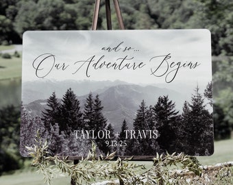May include: A wedding sign with a black and white photo of a mountain range and trees. The text on the sign reads "and so... Our Adventure Begins" with the names "Taylor & Travis" and the date "9.13.25" below.