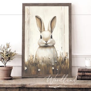May include: A framed print of a rabbit in a field of grass and flowers. The artwork has a rustic, vintage style with a neutral color palette. The frame is a dark brown color. The print is displayed on a wooden table.