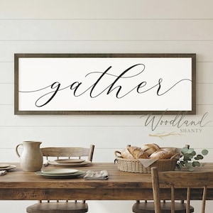 May include: A rustic wooden dining table set for a meal, with a framed sign that reads "gather" in cursive script. The table is surrounded by wooden chairs and set with plates, a pitcher, and a basket of bread.