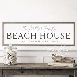 May include: A wooden sign with the text "The Miller Family BEACH HOUSE Myrtle Beach EST. 2026" hangs above a rustic wooden table. A glass jar filled with seashells and a book titled "Coastal Living" are on the table.