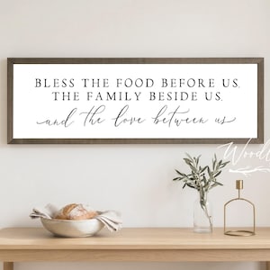 May include: A rectangular wooden sign with the text "BLESS THE FOOD BEFORE US, THE FAMILY BESIDE US, and the love between us" in black script. The sign is above a wooden table with a bowl of bread and a vase.