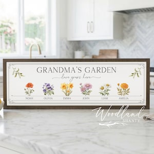 May include: A framed wooden sign with the text "GRANDMA'S GARDEN love grows here." The sign features watercolor illustrations of various flowers, each labeled with a name. The sign is displayed on a white countertop in a kitchen setting.