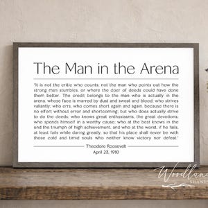 May include: Framed print with the text "The Man in the Arena" by Theodore Roosevelt. The quote is in black font on a white background, framed in a gray wooden frame. A vase of lavender and books are on a wooden surface.