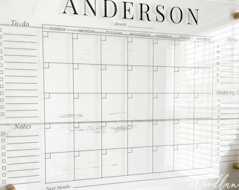 May include: Clear acrylic wall calendar with the name "ANDERSON" at the top. It features a monthly calendar grid, a to-do list, notes section, and shopping list. The calendar is mounted with gold-colored hardware.