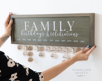 May include: A wooden family birthday and celebrations calendar. The green-painted frame has the words "FAMILY birthdays & celebrations" in white. Wooden discs hang below with names and dates. The calendar is being held up by a person.