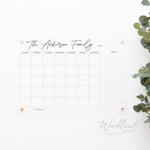 May include: A clear acrylic dry erase calendar with the text "The Anderson Family" at the top. The calendar has a grid for each day of the month, with spaces for notes on the right side. The calendar is mounted on a white wall with gold push pins.