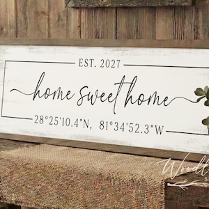 May include: A rustic wooden sign with the words "home sweet home" in elegant script, framed by a black border. The sign includes the text "EST. 2027" and coordinates. The sign is displayed on a wooden bench with a burlap runner.