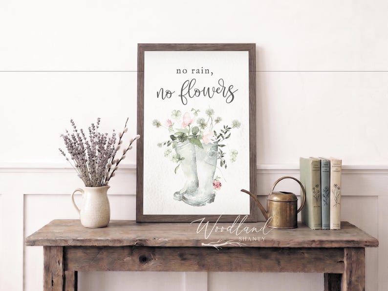 May include: A framed print with the text "no rain, no flowers" above a watercolor image of rain boots filled with flowers. A vase of lavender and books sit on a rustic wooden table.