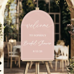 May include: A pink sign on a wooden easel, with the words "Welcome to Sophia's Bridal Shower" and the date 15/08/26. The sign has an arched top and is set against a blurred outdoor background.